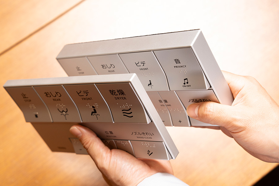 現行品、改良品のウォシュレットリモコンを手に持ち、厚みを比べている。半分以下の薄さになっていることがわかる
