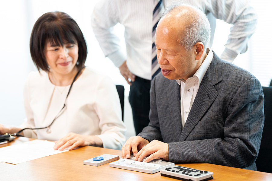 椅子に座り、リモコンを触る長岡さん。西島さんがそれを見ている